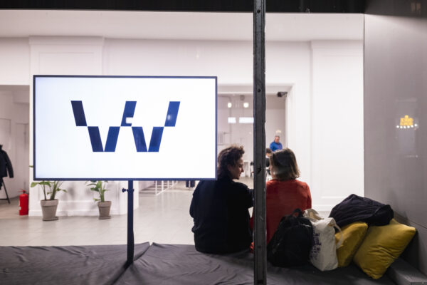 przeszklona witryna okienna WOK Lab, w niej stoi telewizor, na którym wyświetla się ruchome czarne W na białym tle, obok dwie kobiety rozmawiają ze sobą
